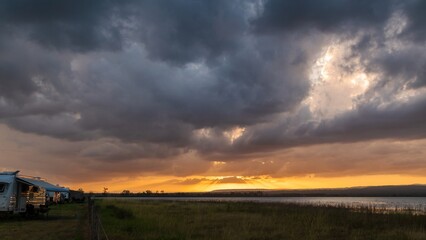 A stunning sunset over Lake Clarendon, Lockyer Valley, Queensland, Australia, with golden sun rays piercing storm clouds, reflecting on the water, and illuminating a peaceful rural camping scene.