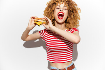 Cheerful young woman with curly hair enjoying a delicious cheeseburger, wearing a red and white striped shirt, against a light background