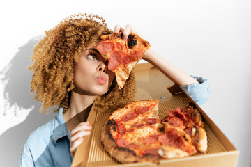 A young woman with curly hair playfully poses with a pizza box, holding a slice of pepperoni pizza and enjoying the moment, showcasing fun, food, and joy
