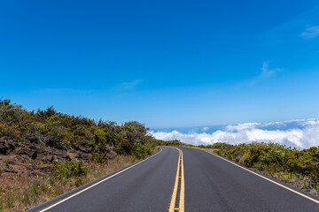 A winding road leads to a horizon of clouds under a clear blue sky.