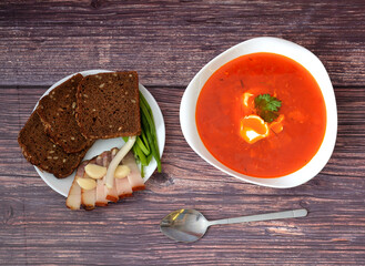 Borscht with sour cream in a deep plate, spoon and dish with rye bread, garlic, lard and green onions on a wooden table.