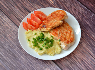 Mashed potatoes with green onions, two pieces of grilled chicken fillet and ripe tomatoes on a wooden table.