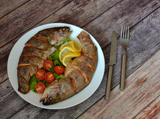 Two whole baked rainbow trout with lettuce, tomatoes and lemon on a wooden table, cutlery nearby.