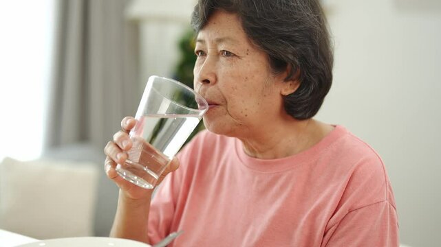 Elderly woman enjoying refreshing water while sitting at a bright table, showcasing the importance of hydration during meals in a cozy home atmosphere