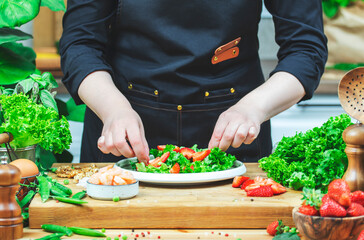 Chef cooking food in the kitchen. Cook prepares gourmet lettuce salad with strawberries and shrimp., wooden table with kitchenware, vegetables, herbs and ingredients. 