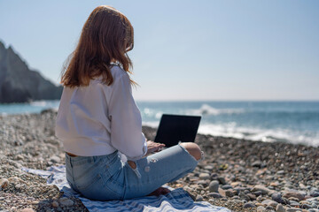 Beach laptop woman: Remote work at sunny beach; Woman using laptop, sitting on blanket by the sea during the day.