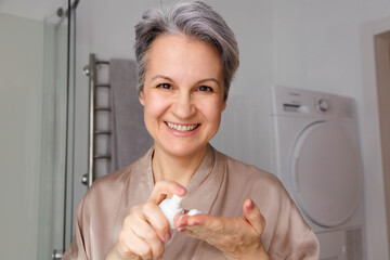 A smiling elderly woman in her age, a pensioner with gray hair, uses a hormonal cream against aging