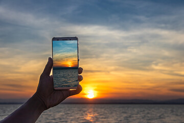 A person capturing a stunning sunrise over the water with a smartphone, highlighting the beauty of nature through modern technology and peaceful morning light.