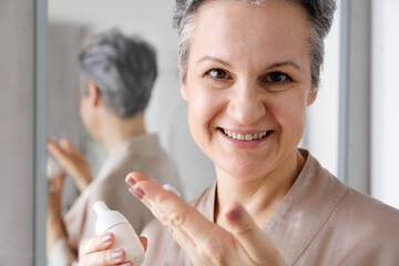 An elderly woman in her age, a pensioner in silk pajamas, takes care of mature skin using a cream