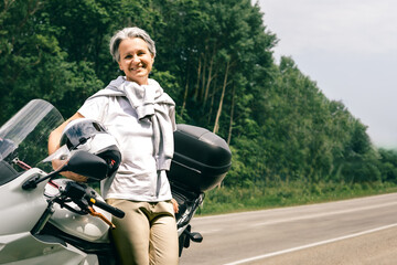 Smiling senior woman with gray hair resting on motorbike by road, celebrating age and freedom