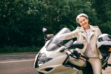 Smiling senior woman with gray hair on motorcycle holding helmet, expressing freedom and confidence