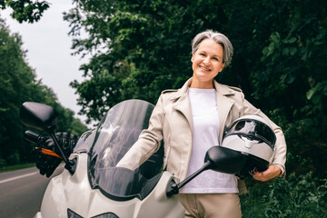 Smiling gray-haired senior woman in beige outfit holding motorcycle helmet on scenic country road