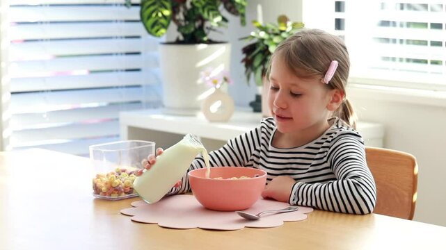 Cute little girl pouring fresh milk from bottle into colorful breakfast cereal. 