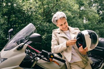 Smiling senior woman with gray hair checking motorcycle helmet, symbol of freedom