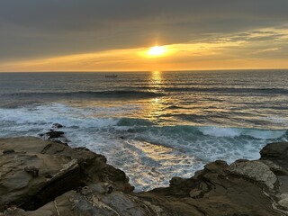 A picturesque sunset over La Jolla, California, featuring a boat on the tranquil water with rugged rocky bluffs framing the foreground, capturing coastal beauty.