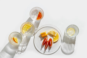 Fruit Infused Water in Glasses and plate with Shadows at sunlight on White background. Aesthetic Top view summer detox drink with slices of lemon and orange with sun flare from glass cups