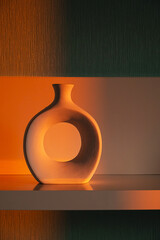 Modern Decorative Ceramic Vase with Circular Hole Stands on Shelf, Illuminated by Warm Orange Light. Home Decor and Style for Interior Design.