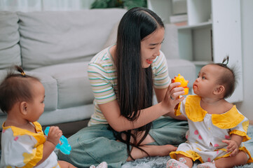 Portrait of Young Asian mother shares a happy moment with her twin daughters while sitting on a soft carpet in a cozy room at home. Leisure activities with family