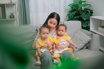 High angle view shot, A young mother sits on a sofa and gently holds her twin daughters in living room at home.