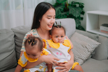 Portrait of Young mother sits on a sofa and gently holds her twin daughters in living room at home.