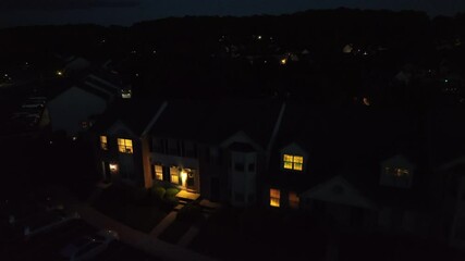 Aerial night view of American suburban neighborhood with glowing house windows and streetlights. Peaceful, cozy atmosphere perfect for themes like home life, evening, or small-town America. Rising.