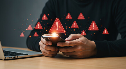 Person using a smartphone with multiple red warning signs floating above it on a desk with a laptop