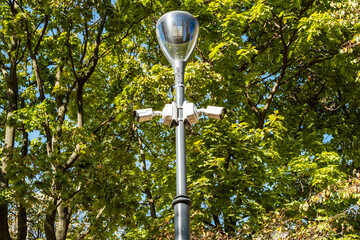 Streetlight pole with multiple security cameras mounted, surrounded by lush green trees. The...