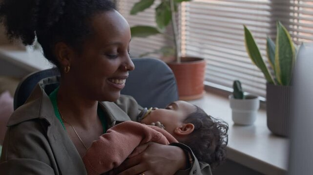 Medium close up shot of cheerful black businesswoman sitting in front of computer with sleeping baby son in arms while having video call in office