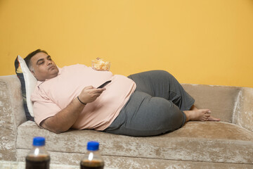 Lazy overweight indian man eating unhealthy food relaxing on sofa at home. Fat guy watch Television...