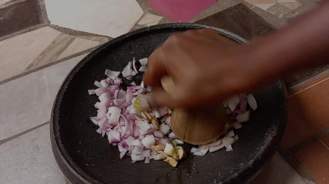 An African grinding pepper, onion, garlic and ginger with a wooden masher in a black earthenware bowl on the floor in the kitchen. Traditional way of preparing food.