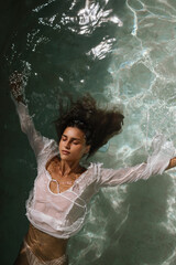 A young woman floats on her back in clear turquoise water, wearing a delicate white shirt. Her face is calm, eyes closed, embodying peace and surrender.