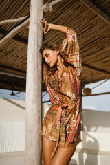 Stylish woman in silk patterned robe posing against a wooden pillar on a sunny tropical terrace. Luxurious bohemian editorial fashion.