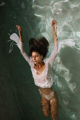 Underwater portrait of a woman floating with arms wide open, dressed in a wet white shirt. The...