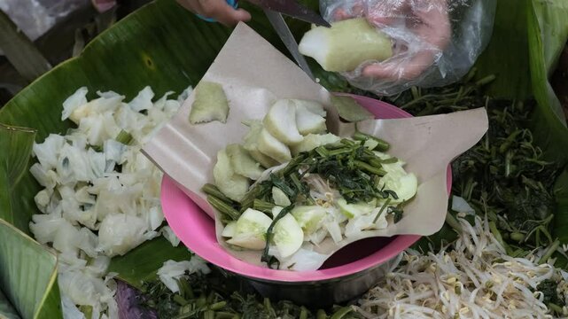 A close-up of a street vendor preparing pecel, a traditional Indonesian salad made from blanched vegetables served with peanut sauce.