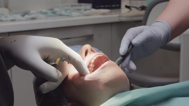 Dental professional works with forceps performing procedure. Assistant manages area with retractor and suction tool ensuring conditions for treatment