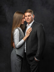 Confident man in business suit standing tall and looking at camera, while graceful woman leans gently against him. Studio portrait on dark background. Elegant and stylish atmosphere