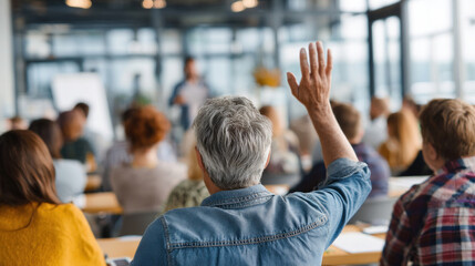 Older male learner with short gray hair raising hand during professional development training, diverse adult students seated around him, bright seminar room