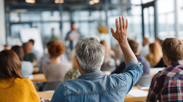 Older male learner with short gray hair raising hand during professional development training, diverse adult students seated around him, bright seminar room