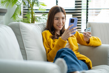 Asian woman shopping online at home with credit card and mobile phone.Happy Asian female sitting on sofa making online payment with credit card and smartphone at home.cashless society concept.