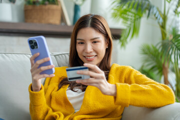 Asian woman shopping online at home with credit card and mobile phone.Happy Asian female sitting on sofa making online payment with credit card and smartphone at home.cashless society concept.
