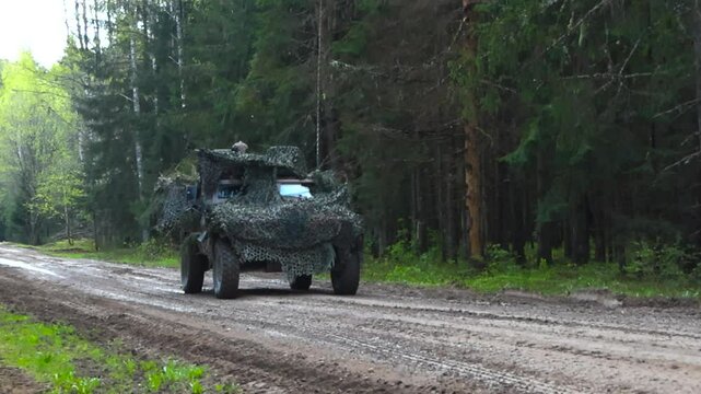 French light armored 4x4 all terrain vehicle or VBL that has a camouflage net on it reversing slowly on a muddy countryside road in a pine forest while two soldiers on top are scouting the enemy area.