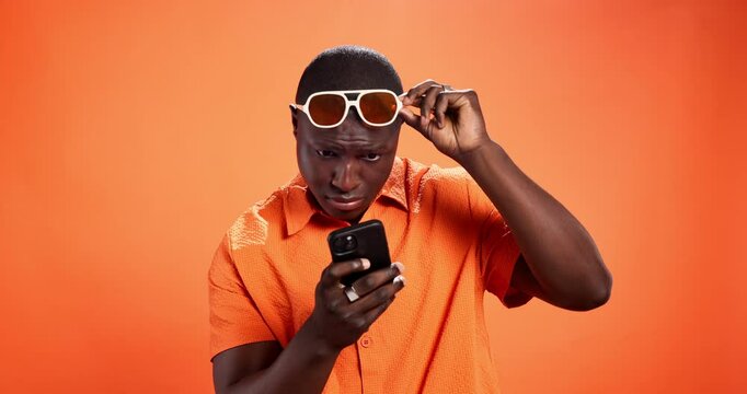 Surprise, smartphone and black man in studio, sunglasses and social media on orange background. African person, wow and model with cellphone, shades and shocked with message and notification with omg