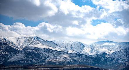 Obraz premium The image shows a majestic mountain range covered in snow under a dramatic sky filled with large, fluffy clouds. The mountains have a rugged and steep appearance, with various peaks and ridges visible