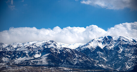The image shows a majestic mountain range covered in snow under a dramatic sky filled with large, fluffy clouds. The mountains have a rugged and steep appearance, with various peaks and ridges visible