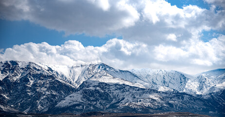 Fototapeta premium The image shows a majestic mountain range covered in snow under a dramatic sky filled with large, fluffy clouds. The mountains have a rugged and steep appearance, with various peaks and ridges visible