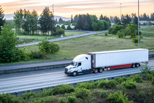 Fototapeta Long haul white big rig semi truck transporting cargo in refrigerator semi trailer driving on the one way highway road at early morning