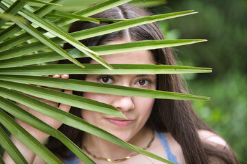 Latin woman, young and beautiful brunette hides behind a large palm leaf showing her face through....