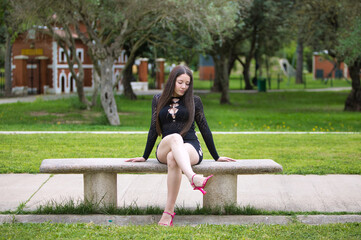 Fototapeta premium Latin woman, young and beautiful brunette dressed in black and pink heels sitting cross-legged on a park bench. The woman makes different expressions and gestures. Olive trees in the background.