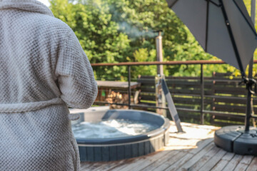 Person in white bathrobe standing near outdoor hot tub with bubbling water on wooden terrace, surrounded by green trees and backyard privacy fence