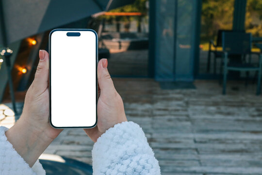 Woman's hands in white robe holding smartphone with blank screen, standing on wooden terrace with stairs, blurred background with modern A-frame cabin and lights, cozy outdoor vacation concept